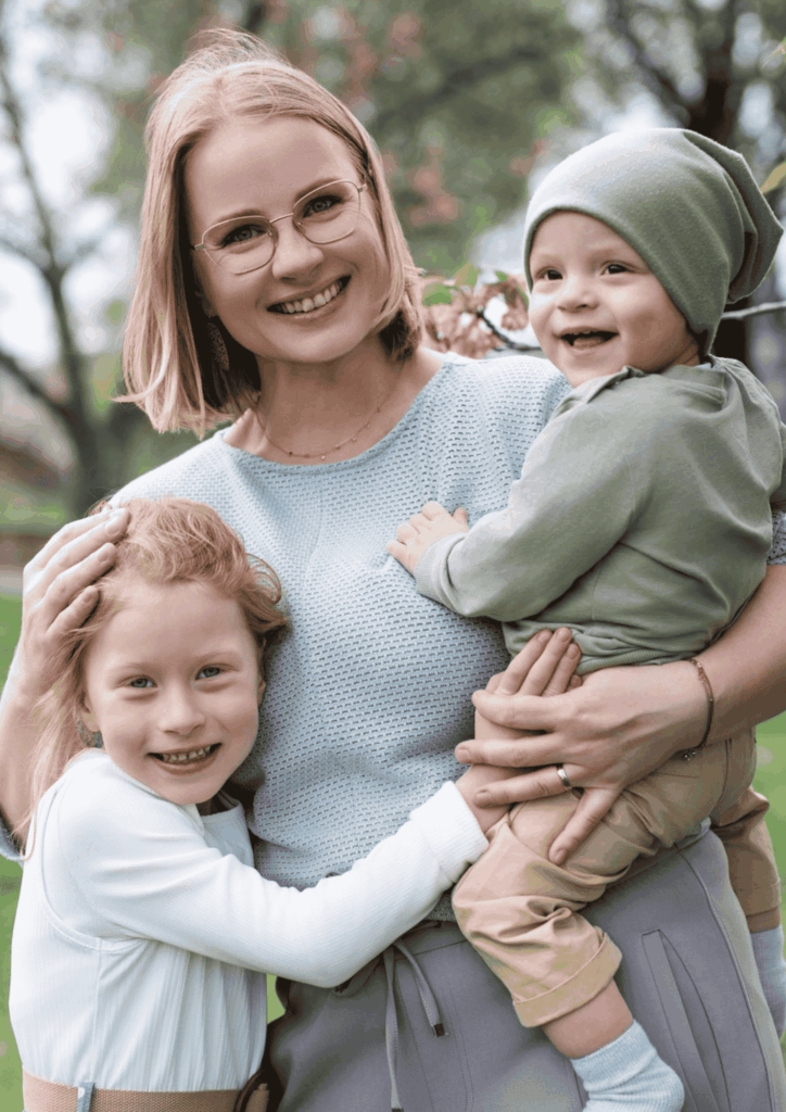 Juliane Krumbiegel lacht gemeinsam mit ihren beiden Kindern im Arm – authentisches Familienportrait im Grünen, Symbol für Mama Mojo, Mental Load Reduktion und Lebensfreude im Mama-Alltag.