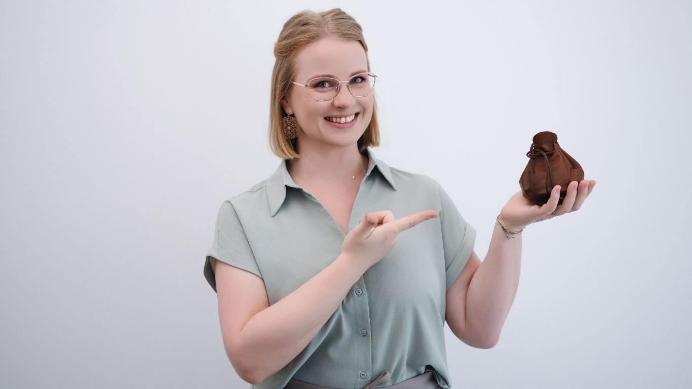 Juliane Krumbiegel, Gründerin von Mama Mojo, lächelt und zeigt auf ein kleines braunes Mojo-Säckchen in ihrer Hand – Symbol für Leichtigkeit, Mental Load Reduktion und Lebensfreude.
