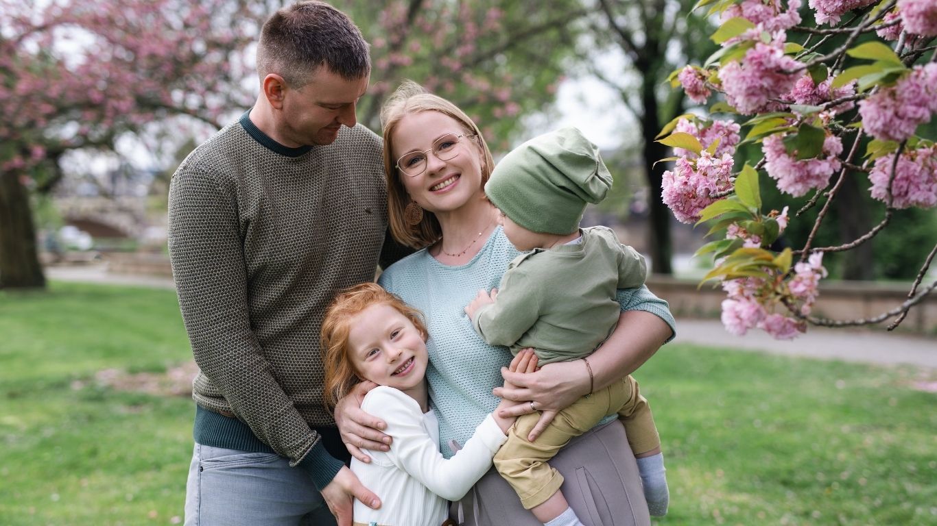 Juliane Krumbiegel lacht gemeinsam mit ihrem Partner und ihren beiden Kindern im Park. Familienfoto mit Kirschblüten im Hintergrund – Symbol für Verbindung, Lebensfreude und Balance im Familienalltag.